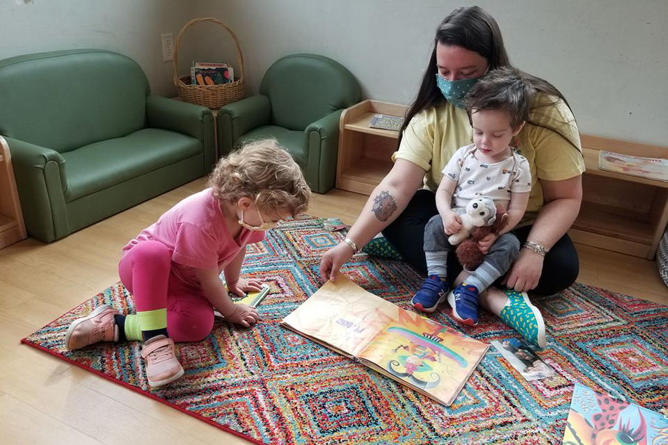 A teacher wearing a mask sits with two children looking at books